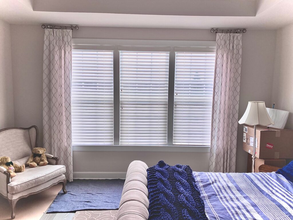 Three tall side by side windows covered by composite blinds in a furnished bedroom with a tray ceiling.