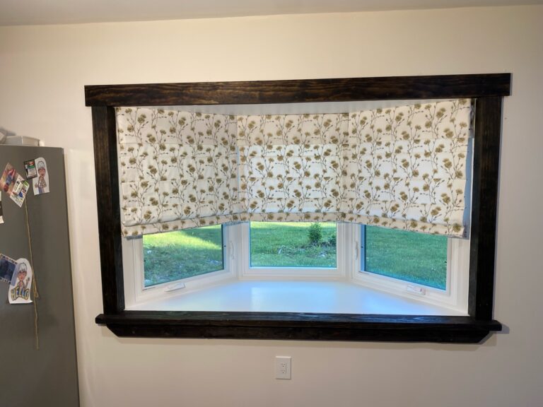 A wide bay window covered by fabric roman shades half opened, allowing light in through the bottom portion.