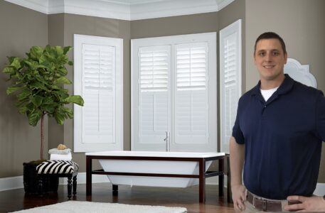 Ryan Bartlett, owner of Bartlett Blinds, standing in front of a bathroom with white plantation shutters surrounding a bathtub.