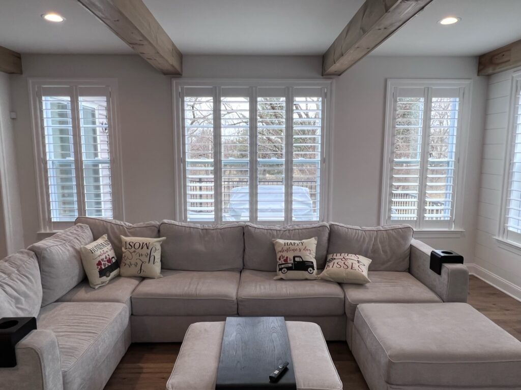 A furnished living room with a large sectional sofa surrounded by Bartlett’s custom interior window shutters.