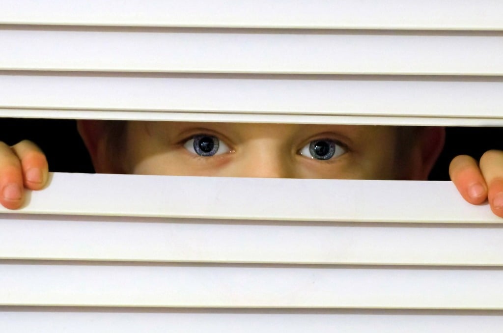 Close-up of a child's eyes peeking through the slats of window blinds. The image highlights the importance of removing hanging cords from blinds to enhance child safety.